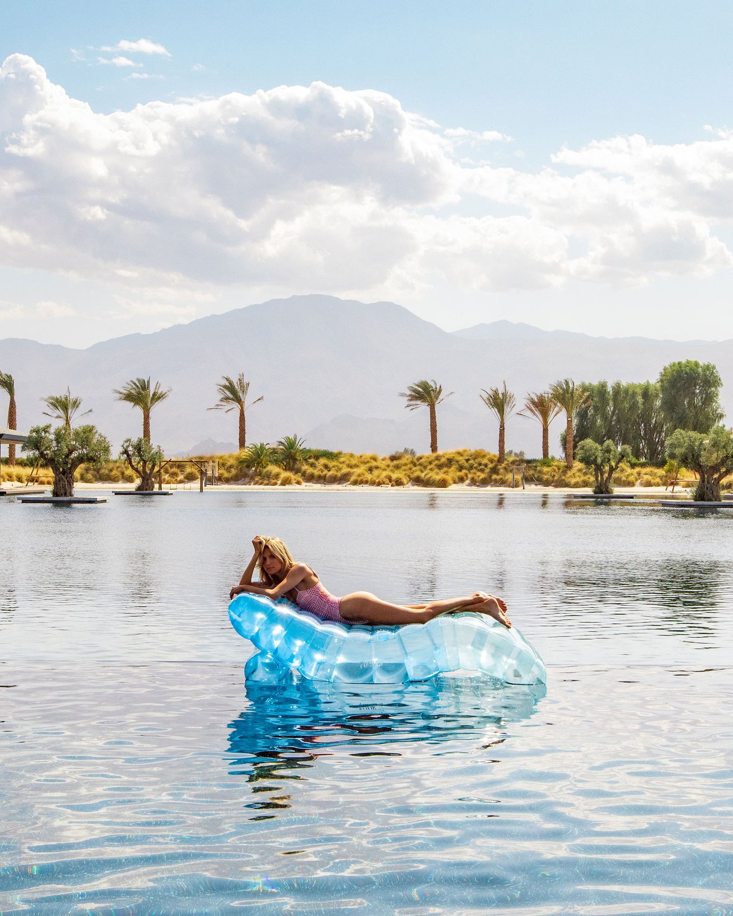 Woman relaxing on a blue FUNBOY lounger float in a serene pool with desert landscape and palm trees in the background.