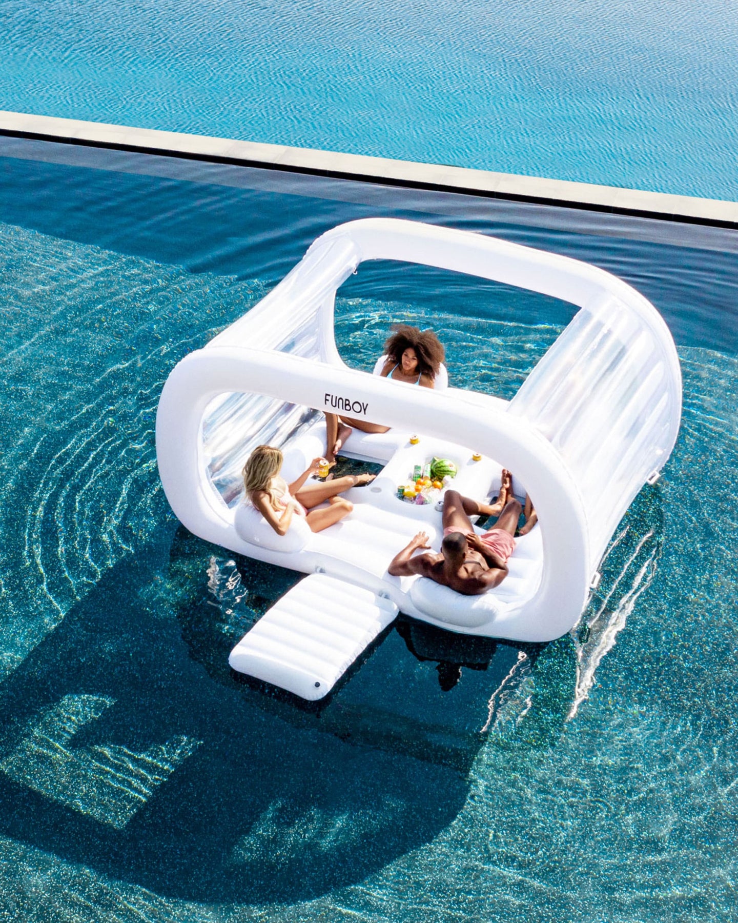 People relaxing on a FUNBOY floating dayclub cabana in a pool, enjoying food and drinks under the sun.