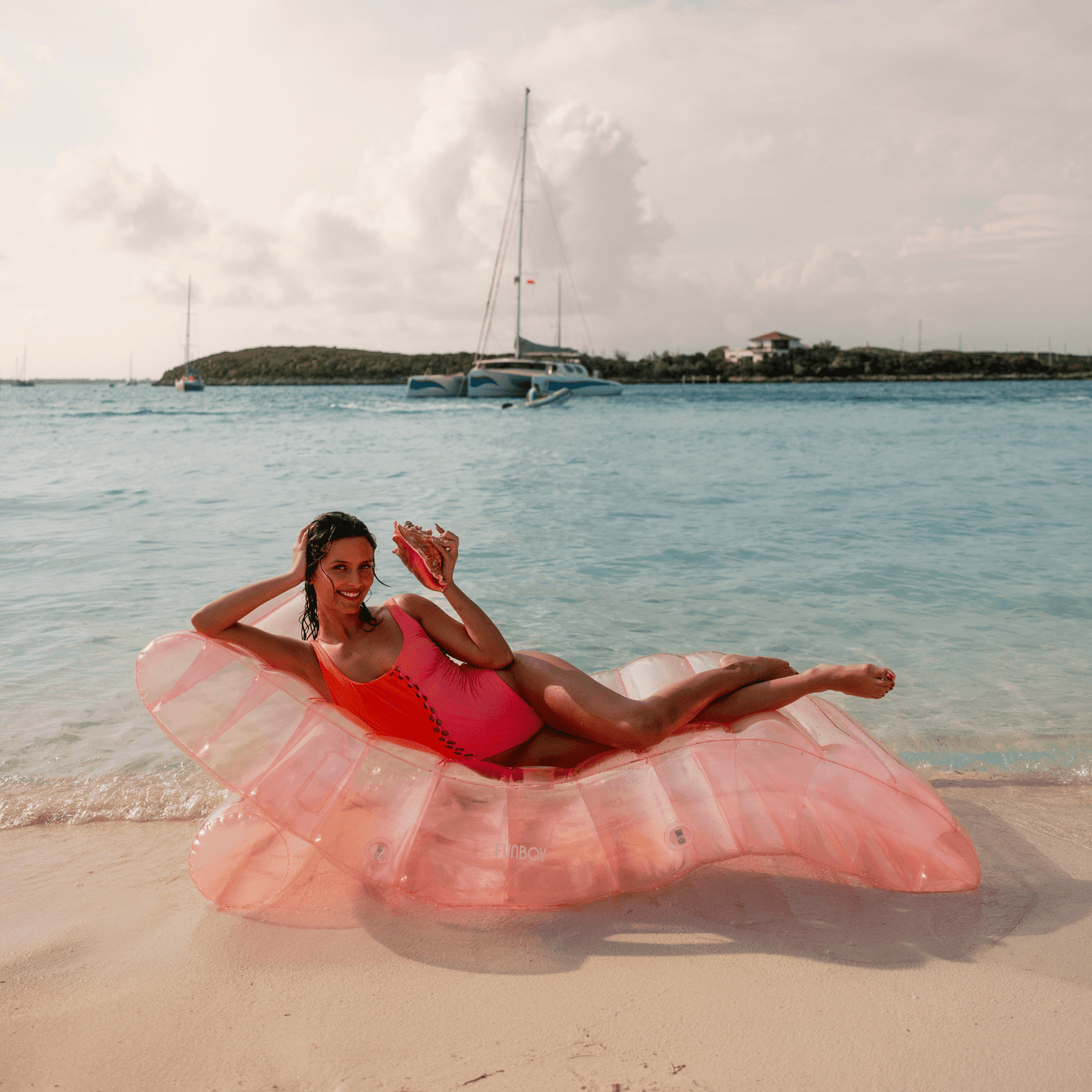 Woman relaxing on a pink FUNBOY lounger pool float at the beach, with a drink in hand and sailboats in the background.